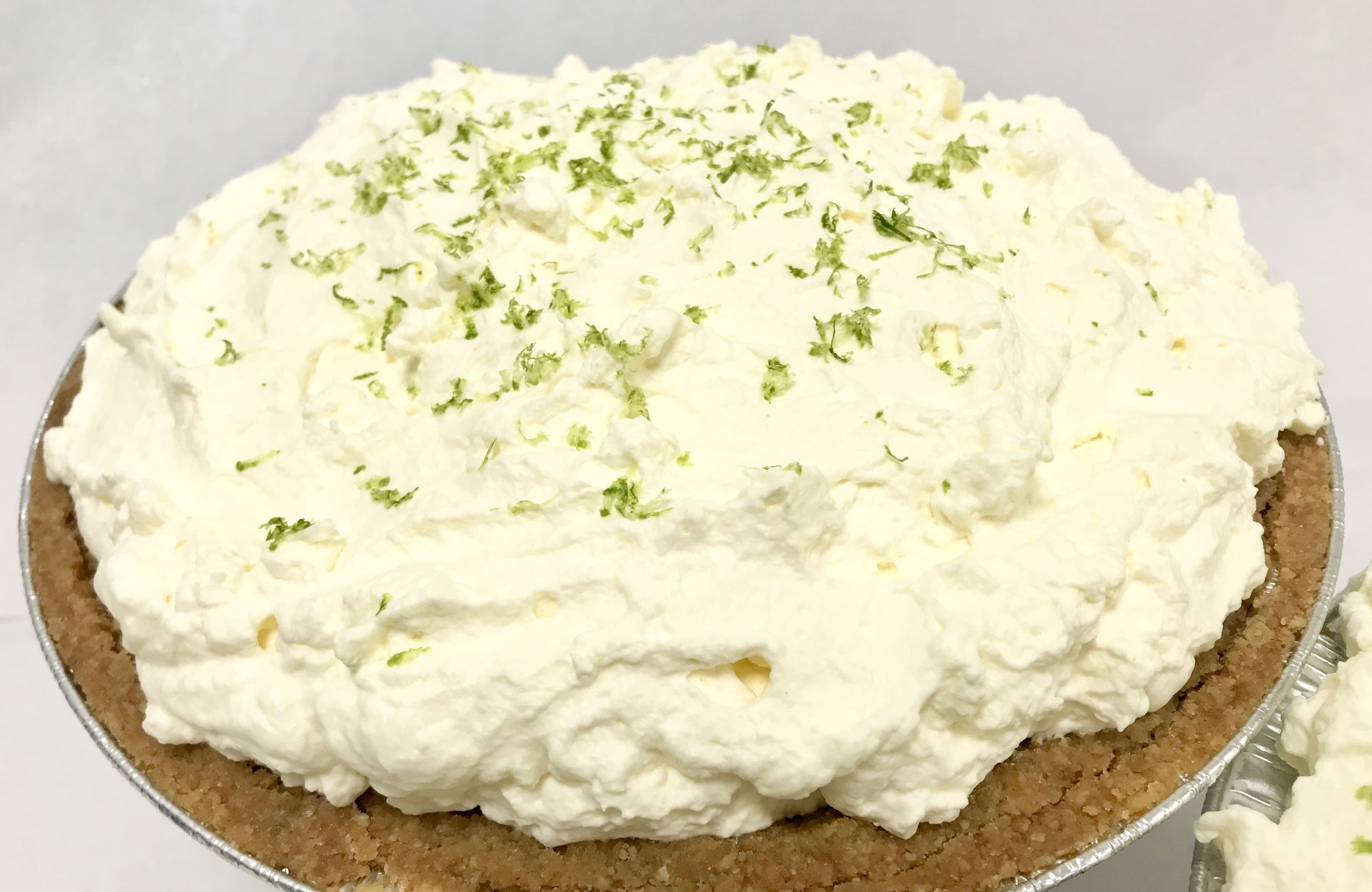 A close-up of a Key Lime Pie with a graham cracker crust, boasting the authentic taste of Key West key limes. The pie is crowned with a generous layer of whipped cream sprinkled with lime zest. The presentation is fluffy and inviting, with a light green garnish highlighting the creamy topping.